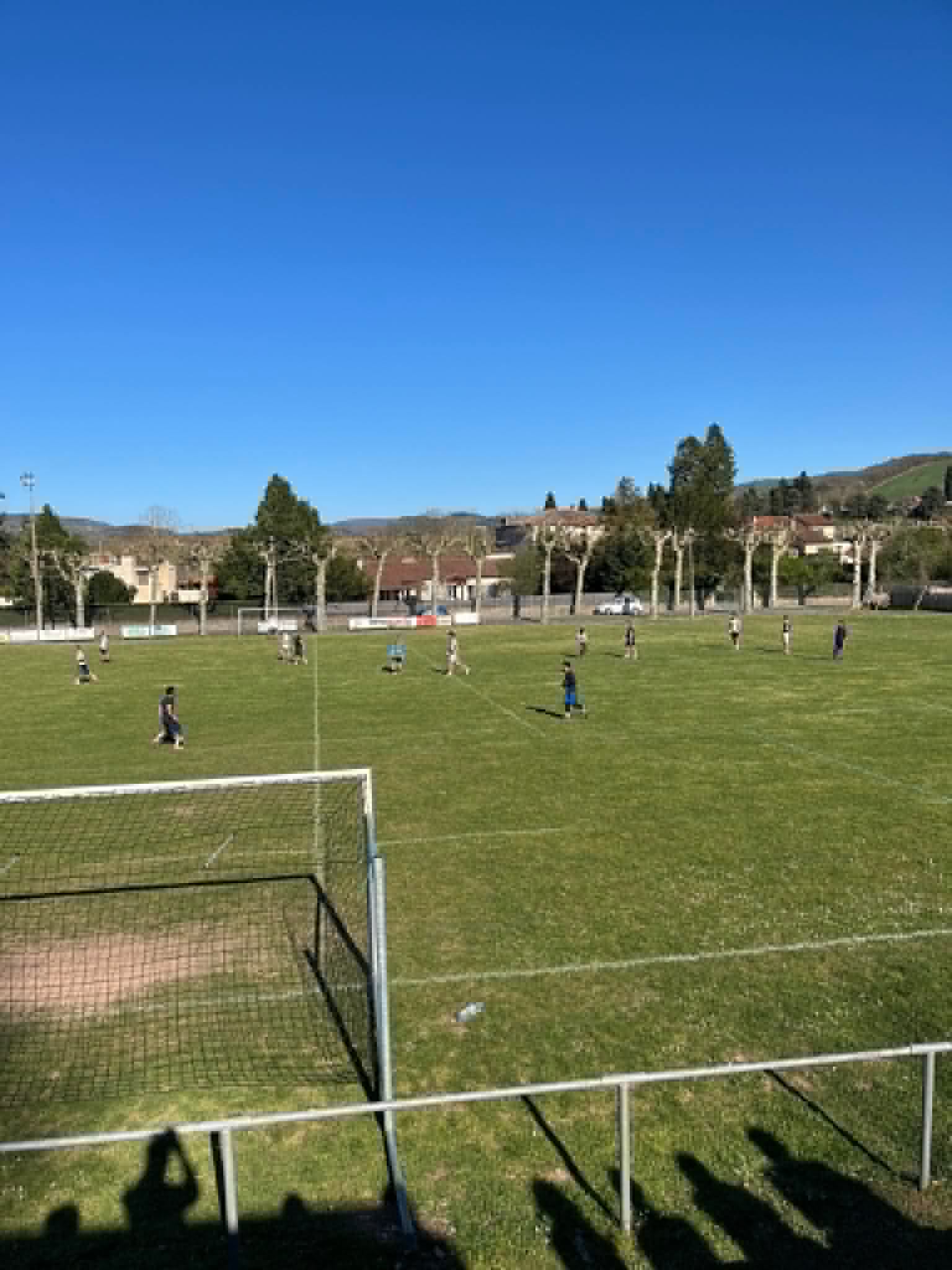 Vue du stade municipal de Camarès pendant le tournoi de Pâques : des équipes jouent sur la pelouse sous un ciel bleu, devant les arbres et les maisons du village.